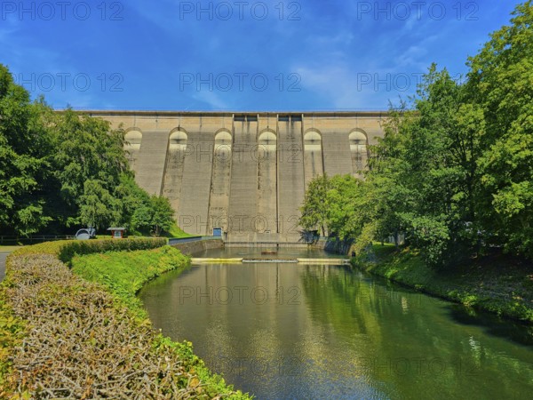Large dam wall in the foreground, flanked by river and lush trees under a clear blue sky,