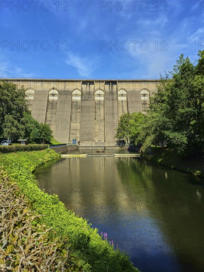 Dam wall with river in the foreground, surrounded by trees and reflected in the water,