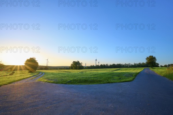 Fork of an asphalt road and a gravel path surrounded by a green meadow with wind turbines in the