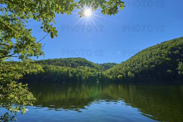 Clear lake landscape with bright sun in the blue sky, surrounded by lush green forests, summer,