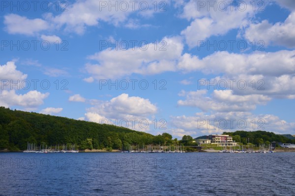 A lake with a building and boats along a wooded shore under a cloudy sky, summer, Rursee,