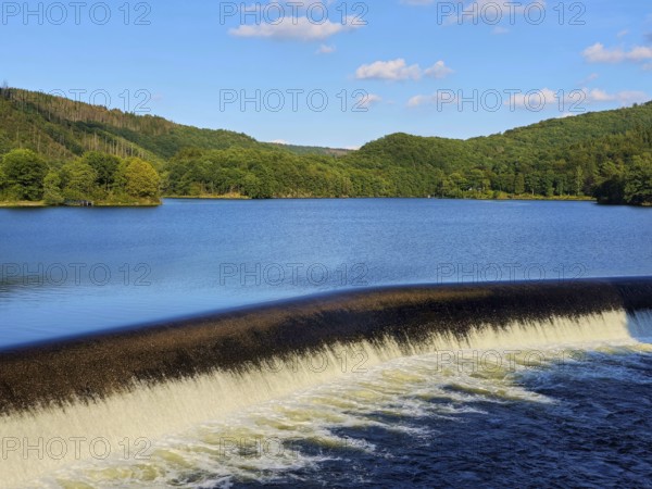 Clear water flows over a dam in a lake surrounded by forests with a blue sky, summer, Rursee,