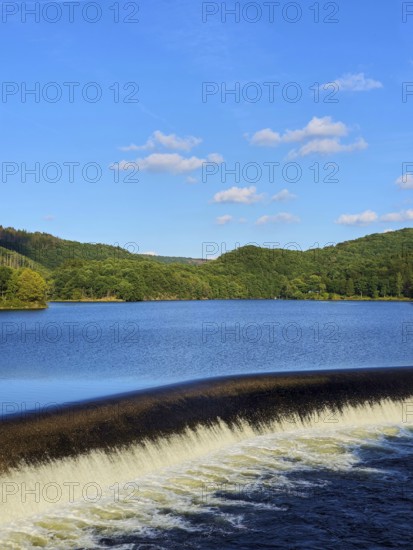 A quiet reservoir with a small waterfall, surrounded by wooded hills under a blue sky, summer,