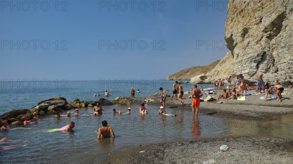 People relaxing on the beach in front of rocky coast and clear blue sea, Embros Thermal Baths, Kos,
