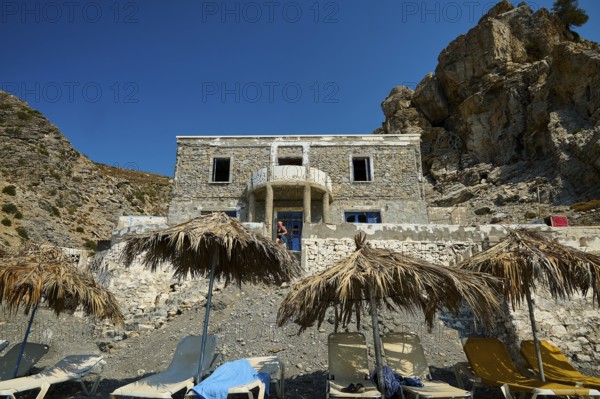 Stone house against a rocky backdrop with beach loungers and thatched roofs in the sunshine, Embros