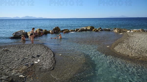 People swimming in the calm blue sea by rocks and sandy shore, Embros Thermal Baths, Kos,