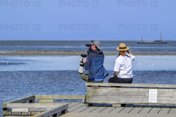 Couple of birdwatchers with binoculars, camera and telephoto lens watching birds at sea from wooden
