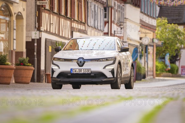 A white electric car drives through a narrow cobbled street with colourful shutters in the town,