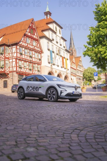 A white car is parked on a cobblestone square next to historic half-timbered buildings with a