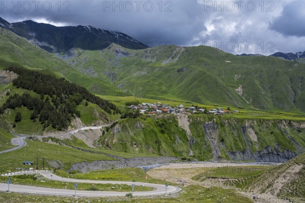 A small village is situated on a hill surrounded by green mountains and cloud-covered sky, views of