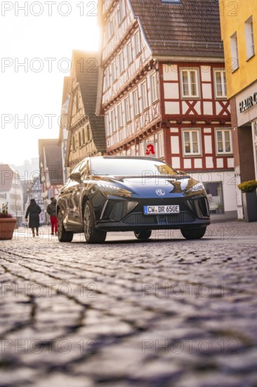 Car drives through an old town with half-timbered houses, some people on foot, MG4 electric car,