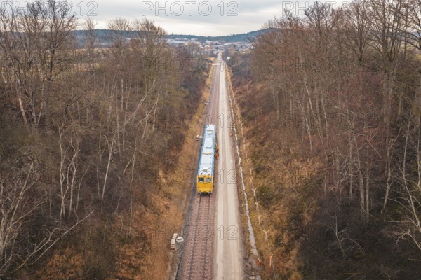 Long view of a rail line with a yellow train through a wintery forest, using a tamping machine at