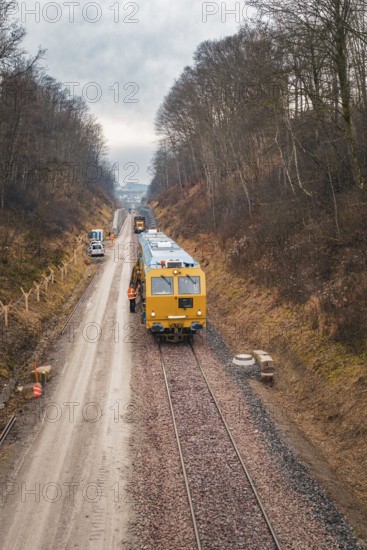 Train with construction workers on a rail line surrounded by winter forest, using a tamping machine