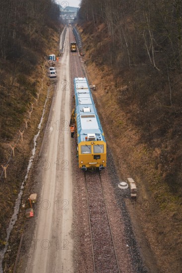 Yellow train on a construction site in a wintry landscape, surrounded by trees, using a tamping