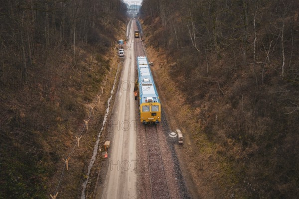 View of a railway line with a yellow train in a lightly wooded area, tamping machine used at the