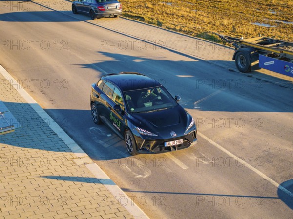 A car is parked on a wide street, illuminated by the evening sun, surrounded by open fields, MG4