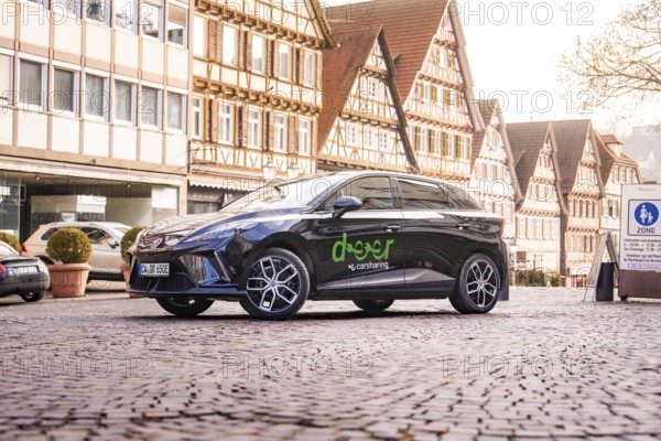 A black car on a cobblestone square surrounded by half-timbered houses in a historic old town, MG4