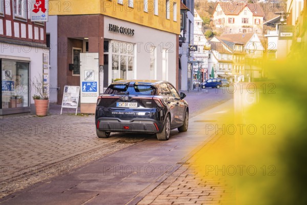 A car drives through a charming shopping street with half-timbered houses and blooming flowers, MG4