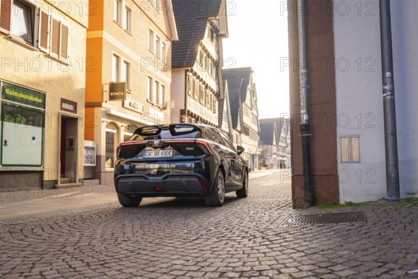 Black car drives through a sunny street lined with historic buildings, MG4 electric car, Deer