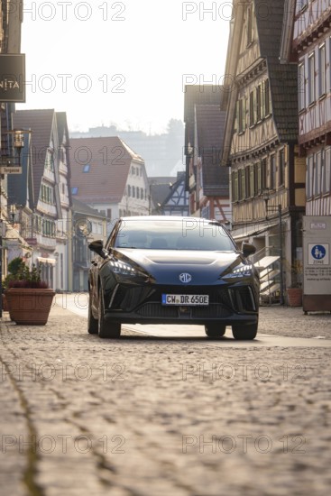 Black car on an empty, paved road surrounded by traditional building facades in sunshine, MG4