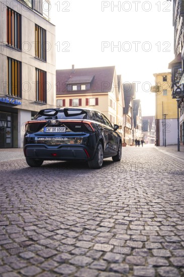 Black car drives through a paved city street between modern and historic buildings in sunlight, MG4