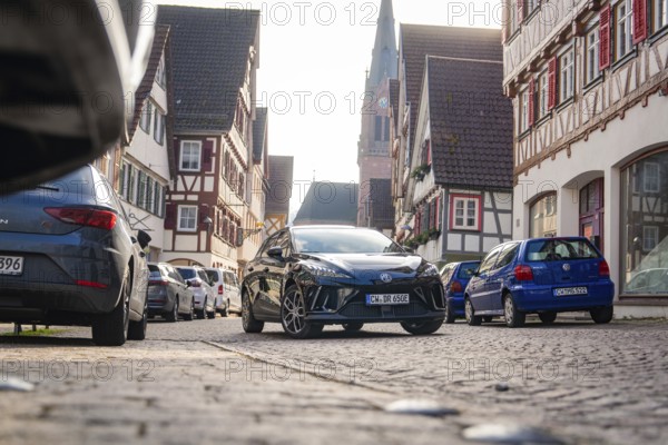 Car in a narrow old town street with half-timbered houses and church in the background, MG4