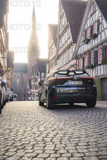 Car drives through a picturesque half-timbered road with church in the background at morning light,