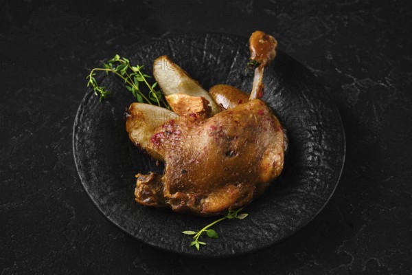 Duck confit on a black stone plate with pieces of roasted pear and herbs. The setting shows a dark