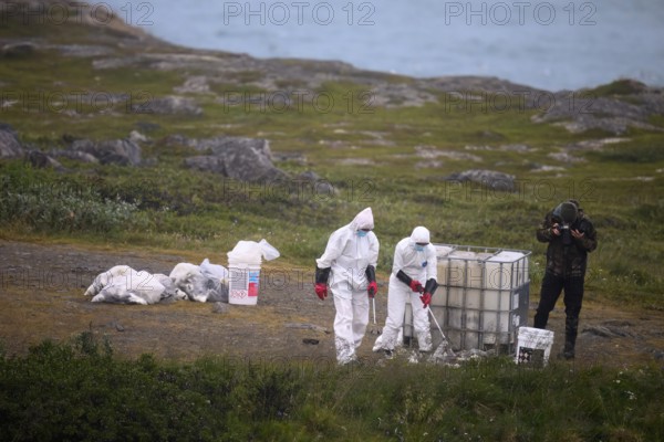 People in protective suits placing dead birds in containers in a coastal landscape, photographer