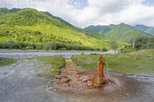 A rusty spring flows onto a gravel riverbed surrounded by green mountains, mineral spring near