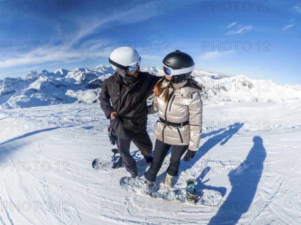 Couple preparing to snowboard on a sunny mountain peak, wearing helmets and goggles with snow