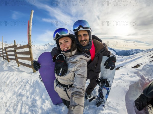 Couple smiling into camera while posing with snowboards on sunny snowy mountain slope, joyful