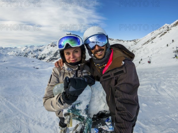 Smiling young couple in winter sportswear and goggles embracing with a snowboard, standing on a