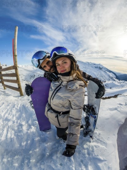 Young couple in helmets and goggles smile at camera, embracing with snowboards on a bright sunny