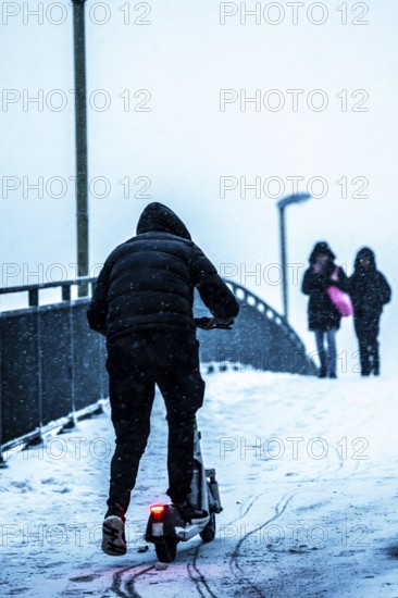Winter weather, blowing snow, man with e-scooter trying to make his way uphill on a snowy sidewalk,