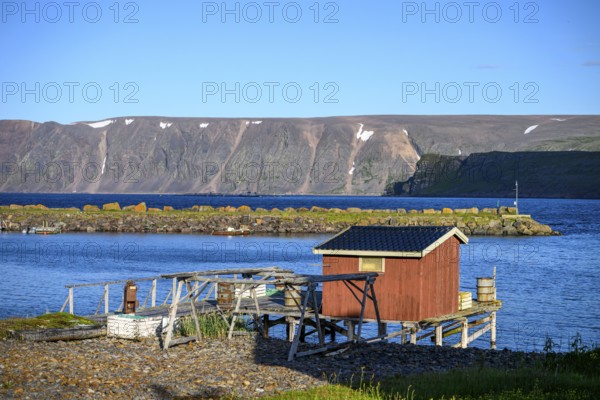 Small wooden cabin on a calm blue sea with mountain backdrop, red house on the shore with a view of