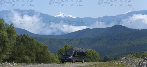 A black van on a road in a mountain valley with snowy peaks and clouds, camper in front of the