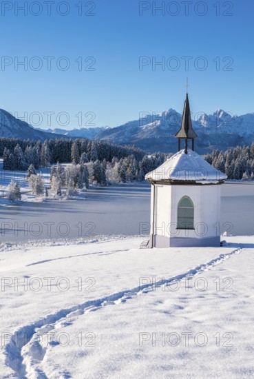 A small chapel in a snow-covered landscape with traces in the snow and mountains in the background,