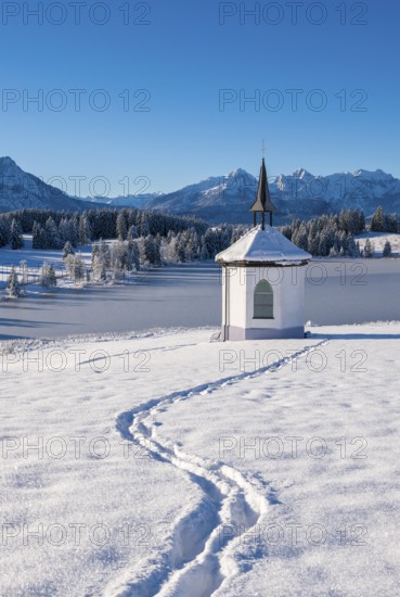 A small chapel in a snowy landscape with mountains in the background and a clear blue sky,