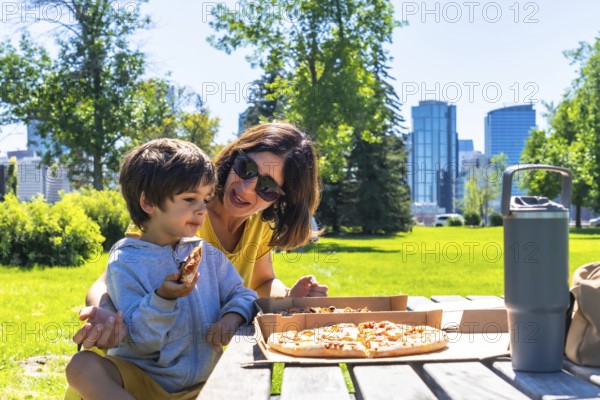 Mother and son are sharing a pizza picnic lunch together on a sunny day at a park in calgary,