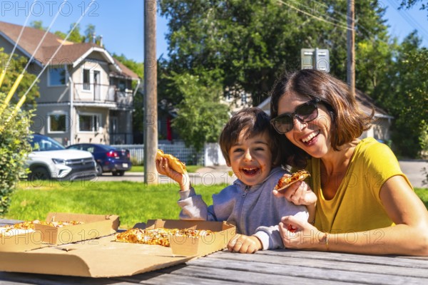 Happy mother and son laughing and eating pizza outdoors at a picnic table in the suburbs of