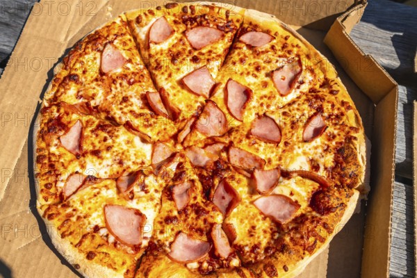 Sliced pizza with ham and melted cheese inside a cardboard box is waiting to be eaten outdoors,