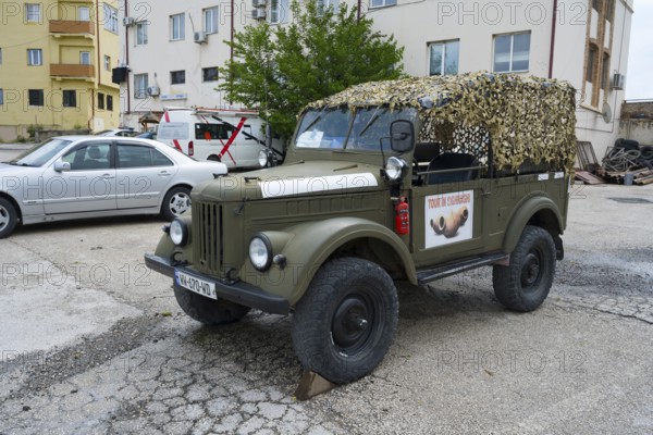 A green vintage car with a camouflage net is parked in a parking lot in front of a building, Soviet