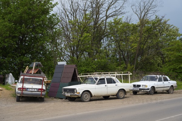 Three older white cars are parked on a rural road next to trees under a cloudy sky, cars with roof