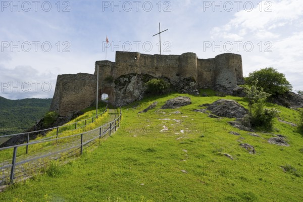 The picture shows medieval castle walls on a hill with surrounding nature under a cloudy sky,