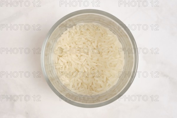 A clear bowl contains uncooked white rice. The rice is fluffy and dry, waiting to be cooked. The
