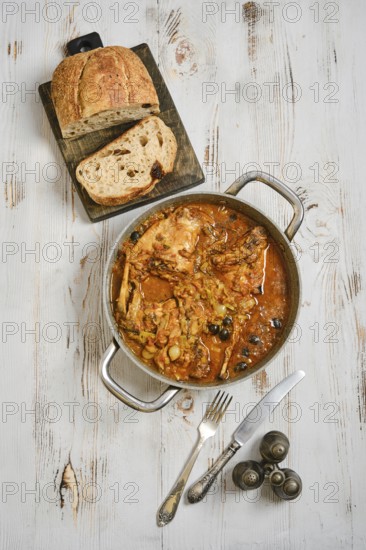 A pot with rabbit stew with vegetables and herbs on a wooden surface. Slices of bread lay on a
