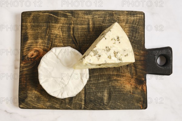 Two types of cheese on a wooden board. One cheese is a wedge with herbs. The other cheese is round
