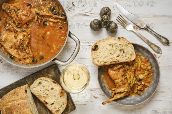 A rabbit stew with olives and vegetables sits in a pan next to a bowl. A piece of bread lies beside