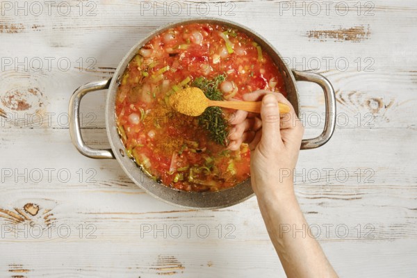 A hand sprinkles spice in a pot with chopped vegetables and a rich tomato base. Spices are added to